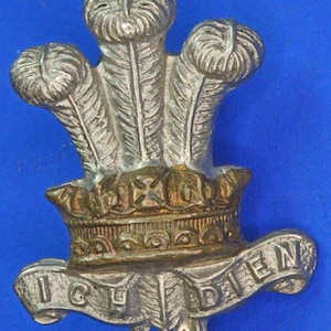 May include: A silver-colored metal badge with three feathered plumes above a crown. The badge has the words "ICH DIEN" on a banner below the crown. The background is a solid blue color.