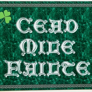 May include: A green metal sign with a Celtic knot border and a shamrock in the top left corner. The sign says "Cead Mile Failte" in white lettering.