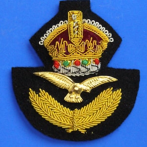 May include: A black felt embroidered patch with a gold crown, eagle, and laurel wreath. The crown has red and green jewels.