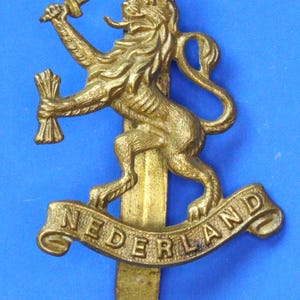 Op de afbeelding: Een messingkleurig embleem met een leeuw die een zwaard en een bundel tarwe vasthoudt. Het woord "NEDERLAND" is gegraveerd op een banner onder de leeuw. Het embleem staat tegen een blauwe achtergrond.