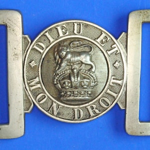 May include: A silver-toned belt buckle featuring the motto "DIEU ET MON DROIT" around a lion and crown emblem. The buckle has two rectangular loops, with a blue background.