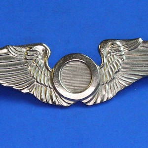 May include: A gold-coloured metal pin featuring a stylised pair of wings flanking a central circular emblem. The wings are detailed with etched feather patterns. The pin is set against a solid blue background.