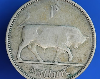 May include: A silver coin featuring a detailed image of a bull walking to the right. The coin has the text "1s" at the top and "Scilling" at the bottom. The coin has a textured edge and is set against a blue background.