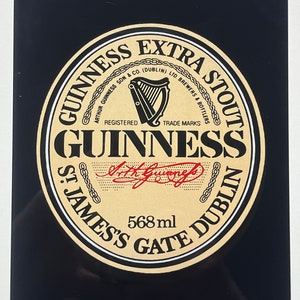May include: A black and gold metal sign with the Guinness Extra Stout logo. The logo features a harp, the words "Guinness Extra Stout", "Arthur Guinness Son & Co. (Dublin) Ltd. Brewers & Bottlers", "Registered Trade Marks", "Guinness", "St. James's Gate Dublin", and "568 ml".