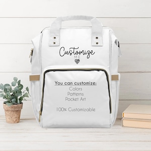 A white diaper travel backpack is displayed upright on a light wood surface with shiplap background and a small potted plant beside it. The backpack reads "Customize Me" in playful script with a heart icon. The front pocket lists customization options: colors, patterns, pocket art, and notes it's "100% Customizable." The aesthetic is bright, white, and airy with a modern farmhouse feel.