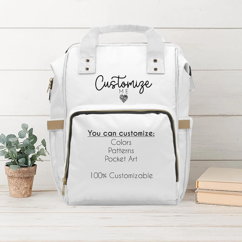 A white diaper travel backpack is displayed upright on a light wood surface with shiplap background and a small potted plant beside it. The backpack reads "Customize Me" in playful script with a heart icon. The front pocket lists customization options: colors, patterns, pocket art, and notes it's "100% Customizable." The aesthetic is bright, white, and airy with a modern farmhouse feel.