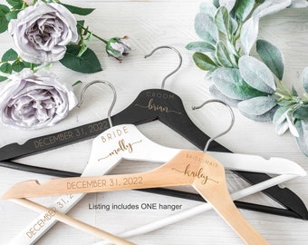 May include: Three wooden hangers with personalized text. The top hanger is black with the text "GROOM brian" and "DECEMBER 31, 2022". The middle hanger is white with the text "BRIDE molly" and "DECEMBER 31, 2022". The bottom hanger is natural wood with the text "BRIDESMAID kailey" and "DECEMBER 31, 2022". The text "Listing includes ONE hanger" is also visible.