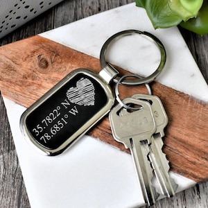 May include: A rectangular silver keyring with a black centre featuring a heart design and coordinates. The keyring is attached to a silver ring with two keys. The coordinates are 35.7822° N 78.6851° W.
