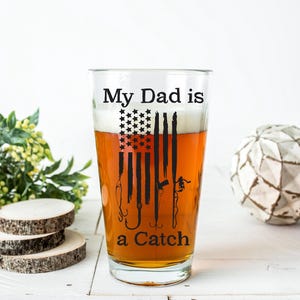 May include: A clear pint glass filled with beer, featuring a black graphic of the American flag with fishing hooks and the text "My Dad is a Catch." The glass is on a white surface with decorative elements.