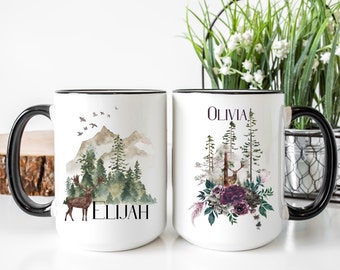 May include: Two white ceramic mugs with black rims and handles. One mug features a watercolor design of a deer, trees, mountains, and the name "ELIJAH". The other mug has a similar design with the name "OLIVIA" and floral accents.