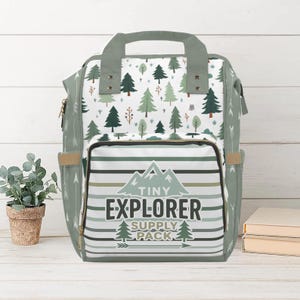 以下が含まれることがあります： 森のパターンと「Tiny Explorer Supply Pack」の文字が入った、緑と白のバックパック。 バックパックには上部のハンドル、サイドポケット、水平ストライプのフロントポケットが付いています。 デザインには木と山が含まれています。