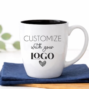Custom logo coffee mug in classic white ceramic, 18 oz glossy bistro mug with black interior and permanent laser engraved branding. This timeless two-tone ceramic cup features a smooth curved handle and professional etched finish, photographed in a bright white farmhouse kitchen—ideal branded coffee mug for small business marketing, corporate gifts, client appreciation, or employee welcome kits.
