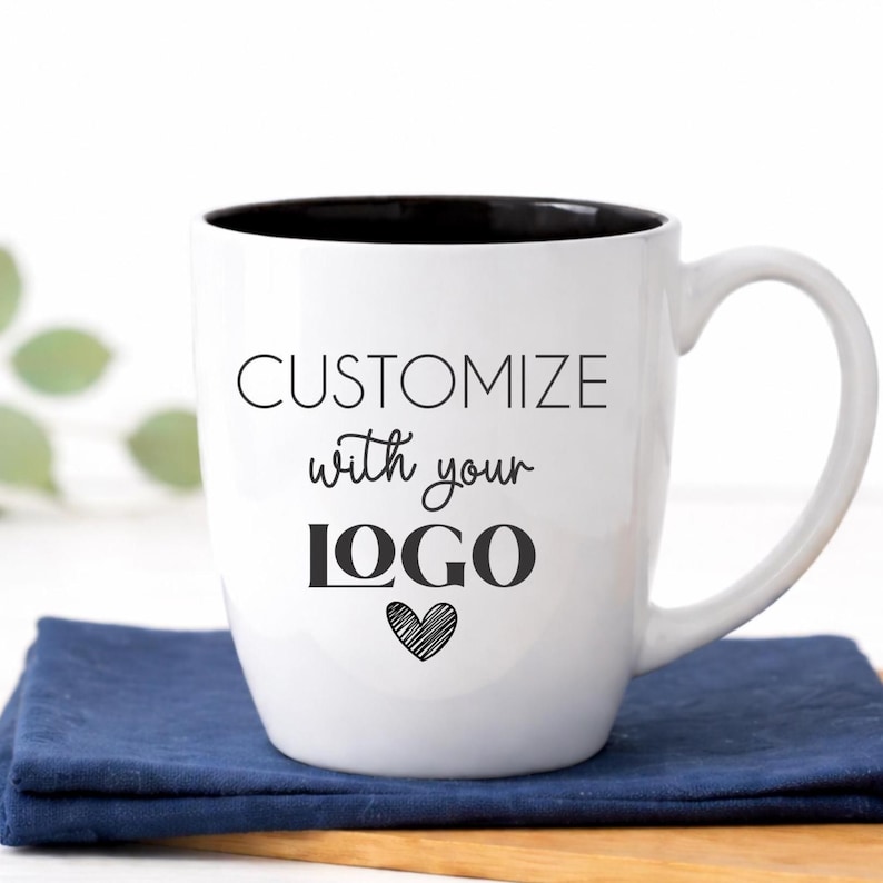 Custom logo coffee mug in classic white ceramic, 18 oz glossy bistro mug with black interior and permanent laser engraved branding. This timeless two-tone ceramic cup features a smooth curved handle and professional etched finish, photographed in a bright white farmhouse kitchen—ideal branded coffee mug for small business marketing, corporate gifts, client appreciation, or employee welcome kits.