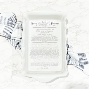 Image of a white ceramic ruffled casserole dish engraved with a recipe titled “Granny’s Piggies.” The design includes a playful pig illustration, a list of ingredients, and detailed instructions in a mix of print and script fonts. The dish is placed on a marble surface with a blue and white checkered towel.