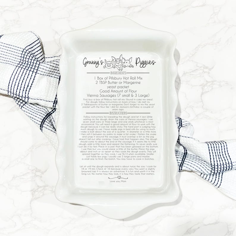 Image of a white ceramic ruffled casserole dish engraved with a recipe titled “Granny’s Piggies.” The design includes a playful pig illustration, a list of ingredients, and detailed instructions in a mix of print and script fonts. The dish is placed on a marble surface with a blue and white checkered towel.