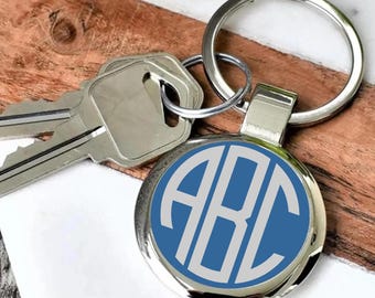 May include: A silver keychain with a round, blue and white monogram design featuring the letters "ABC". The keychain is attached to a silver key ring and a set of keys. The background is a wood and marble surface.