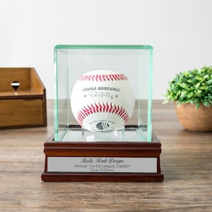 May include: A wooden display case with a glass top and a baseball inside. The baseball is white with red stitching and has the text "USSSA Baseball" printed on it. The case has a silver plaque with the text "Rustic Route Designs Annual Co-Ed League Captain Ryan Williams".