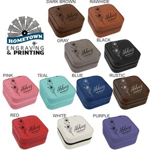 A collection of personalized travel jewelry cases is displayed in various colors, including dark brown, rawhide, gray, black, pink, teal, blue, rustic, red, white, and purple. Each case features an engraved floral design with a customizable name or text, making them ideal for bridesmaid gifts or personalized keepsakes. The Hometown Engraving & Printing logo is visible, indicating customization services.