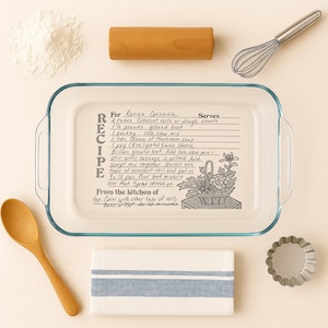 May include: A clear glass baking dish with a handwritten recipe for Runza Casserole, surrounded by a wooden rolling pin, whisk, flour, a wooden spoon, a striped towel, and a small metal tart pan. The recipe includes ingredients and baking instructions.