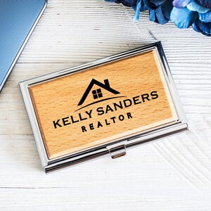 K&ouml;nnte beinhalten: Ein Visitenkartenhalter aus Holz mit einem silbernen Metallrahmen. Das Holz ist hellbraun und das Metall gl&auml;nzend. Der Halter hat ein Haussymbol mit einem Fenster und den Text "Kelly Sanders REALTOR" in das Holz graviert.