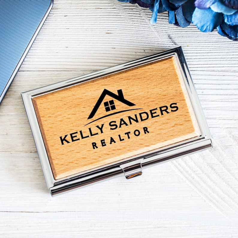 K&ouml;nnte beinhalten: Ein Visitenkartenhalter aus Holz mit einem silbernen Metallrahmen. Das Holz ist hellbraun und das Metall gl&auml;nzend. Der Halter hat ein Haussymbol mit einem Fenster und den Text "Kelly Sanders REALTOR" in das Holz graviert.