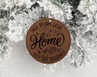 May include: Brown leather ornament with the text "All roads lead home for the holidays" in black lettering. The ornament features a small house illustration and snowflakes.