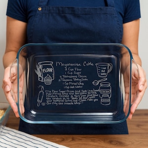 May include: A clear rectangular glass baking dish with handles, held by a person. The dish has a chalkboard-style recipe for "Mayonnaise Cake" written in white. The recipe includes ingredients like flour, sugar, cocoa, and baking soda.