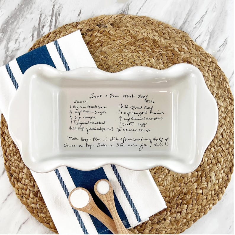 White ceramic loaf pan with a laser-engraved handwritten recipe for Sweet and Sour Meat Loaf, featuring ingredients and instructions. The pan is placed on a woven placemat with a blue and white striped kitchen towel and wooden measuring spoons. A personalized and functional keepsake, perfect for preserving cherished family recipes or gifting.