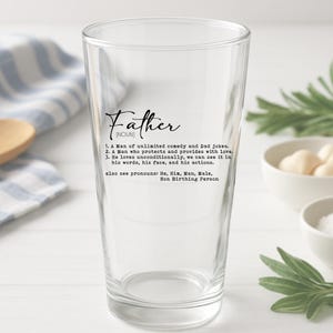 May include: A clear pint glass with the word "Father" in a script font. Below the word is a definition of "Father" including the phrases "unlimited comedy and Dad jokes" and "Non Birthing Person".