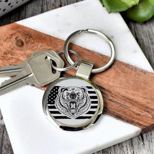 May include: A round silver keychain with a black and white graphic of a bear's head over an American flag design. The keychain is attached to a silver key ring. Keys are visible in the background.