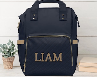 May include: Navy blue backpack with a quilted front pocket. The pocket features the name "LIAM" in gold lettering. The bag has a top handle, gold zippers, and tan accents. A small potted plant is visible on the left.