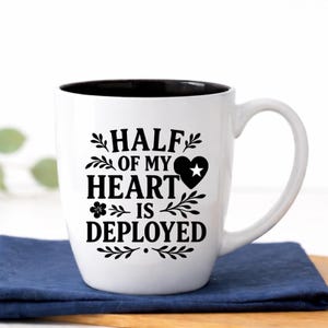 Half of My Heart Is Deployed military family coffee mug in classic white ceramic, 18 oz glossy bistro cup with black interior and precision laser engraved patriotic design. This sturdy two-tone ceramic mug features elegant leafy accents and heart star detail, photographed in a bright farmhouse setting. Thoughtful gift for military spouse, deployment keepsake, National Guard or active duty family gift, welcome home surprise, or supportive patriotic present.