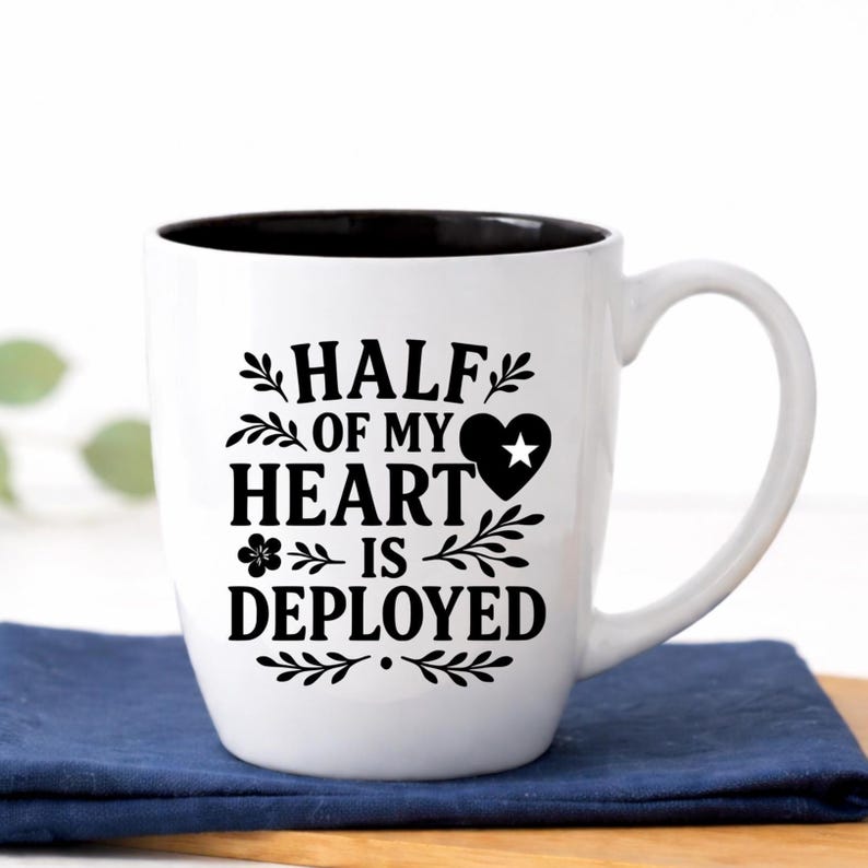 Half of My Heart Is Deployed military family coffee mug in classic white ceramic, 18 oz glossy bistro cup with black interior and precision laser engraved patriotic design. This sturdy two-tone ceramic mug features elegant leafy accents and heart star detail, photographed in a bright farmhouse setting. Thoughtful gift for military spouse, deployment keepsake, National Guard or active duty family gift, welcome home surprise, or supportive patriotic present.