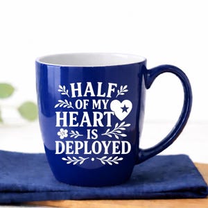 Half of My Heart Is Deployed military spouse coffee mug in glossy royal blue ceramic, 18 oz bistro style cup with white interior and permanent laser engraved deployment quote. This durable two-tone ceramic mug features bold farmhouse typography with heart and star detail, curved handle, and high-shine finish. Styled in a bright white airy kitchen with navy linen and wood board—perfect military wife gift, deployment gift for her, Army Navy Air Force support mug, or patriotic care package idea.
