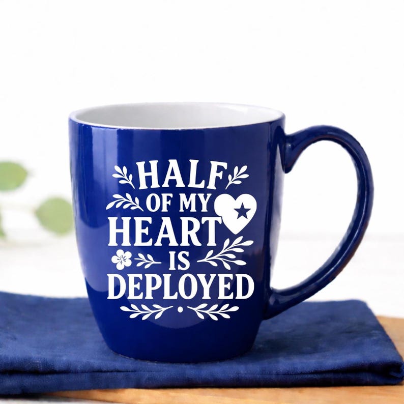 Half of My Heart Is Deployed military spouse coffee mug in glossy royal blue ceramic, 18 oz bistro style cup with white interior and permanent laser engraved deployment quote. This durable two-tone ceramic mug features bold farmhouse typography with heart and star detail, curved handle, and high-shine finish. Styled in a bright white airy kitchen with navy linen and wood board—perfect military wife gift, deployment gift for her, Army Navy Air Force support mug, or patriotic care package idea.