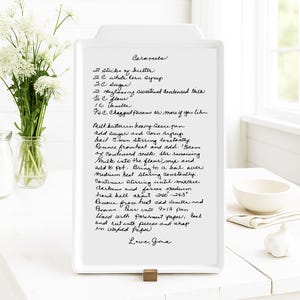 May include: A white ceramic recipe board with a handwritten caramel recipe. The recipe includes ingredients like butter, sugar, and pecans. The board is propped up on a wooden stand, with a vase of white flowers and a bowl in the background.