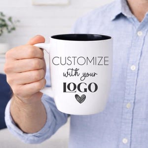 Custom logo bistro mug in hand, 18 oz glossy white ceramic coffee cup with black interior, professionally laser engraved for permanent, high-contrast branding. This large personalized business mug is perfect for corporate gifts, employee appreciation, client thank you gifts, real estate closing gifts, and small business merchandise. Shown held for scale in a bright farmhouse setting with soft natural light.