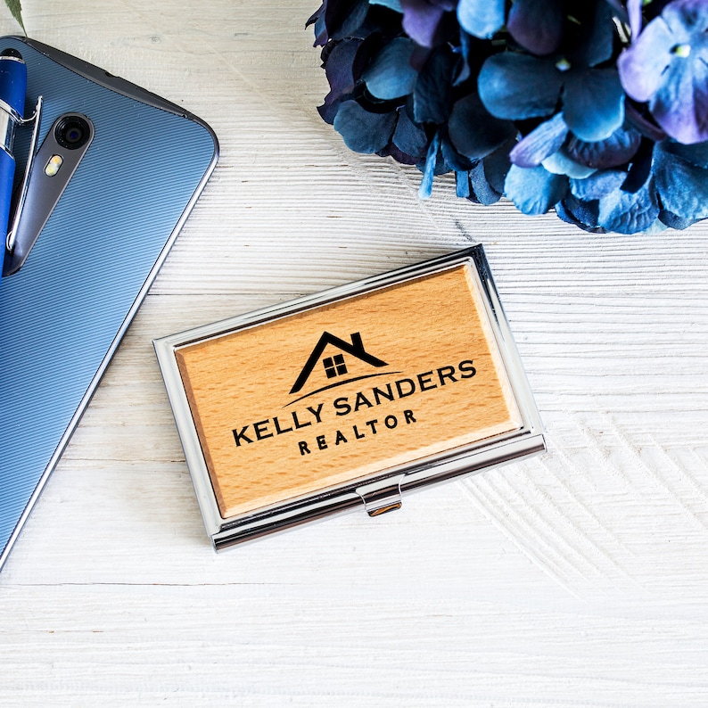 K&ouml;nnte beinhalten: Ein Visitenkartenhalter aus Holz mit einem silbernen Metallrahmen. Der Halter hat ein schwarz-wei&szlig;es Logo mit einem Haus und dem Text "Kelly Sanders REALTOR".