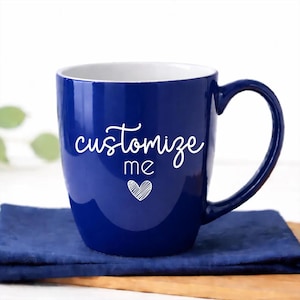 May include: A deep blue ceramic mug with a white interior. The mug features the words "customize me" in a cursive font, with a heart graphic below. The mug is on a blue napkin.