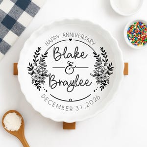 May include: White ceramic pie dish with a fluted edge, featuring the text "Happy Anniversary" and names "Blake & Braylee" with a floral wreath design. The date "December 31, 2026" is also included. The dish rests on a wooden stand.
