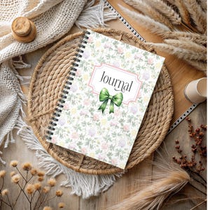 May include: A spiral-bound journal with a floral pattern cover. The cover features a pink-framed label with the word "Journal" and a green bow. The background is a mix of pastel flowers and green vines. The journal rests on a woven tray.