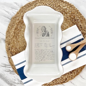 May include: White ceramic loaf pan with a scalloped edge. The pan features a black and white illustration of a woman with a handwritten recipe for "Grammy's Famous Date Nut Bread".