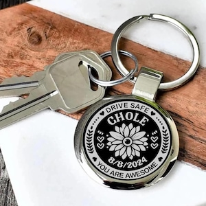 May include: A silver-colored keychain with a round pendant featuring the name "CHOLE" in bold letters, a floral design, and the date "8/8/2024". The keychain also includes the text "DRIVE SAFE" and "YOU ARE AWESOME". Keys are visible.