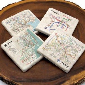 May include: Four square coasters with city map designs, including Kyoto, Miami, and Paris, are arranged on a wooden surface. The coasters feature colorful lines representing transportation routes and city names.