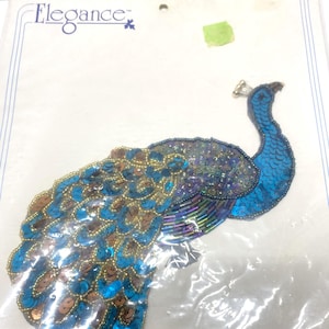 May include: A sequined peacock applique in a plastic package. The peacock features blue, teal, and gold sequins, with a detailed design. The word "Elegance" is printed at the top left corner of the package.
