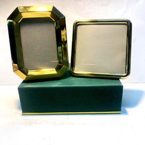 May include: Two gold-toned metal picture frames with a polished finish. One frame is rectangular with beveled edges, and the other is square with rounded corners. Both frames feature light-colored matting and are displayed on a dark green box.