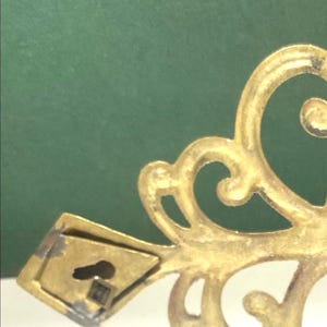 Puede incluir: Primer plano de una pieza decorativa de metal dorado y ornamentada. El dise&ntilde;o presenta patrones arremolinados en forma de volutas y un peque&ntilde;o soporte de montaje rectangular con un ojo de cerradura. El fondo es verde liso y apagado.