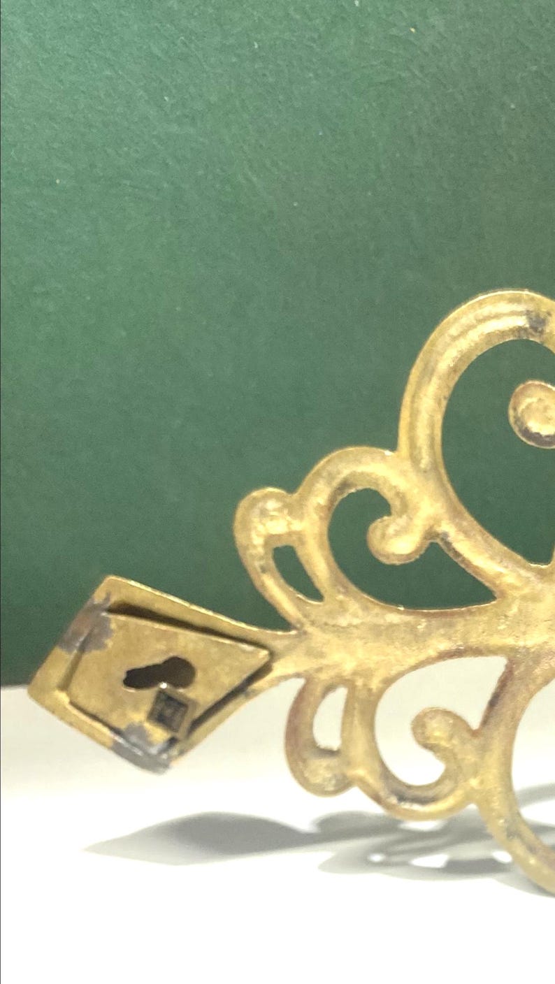 Puede incluir: Primer plano de una pieza decorativa de metal dorado y ornamentada. El dise&ntilde;o presenta patrones arremolinados en forma de volutas y un peque&ntilde;o soporte de montaje rectangular con un ojo de cerradura. El fondo es verde liso y apagado.