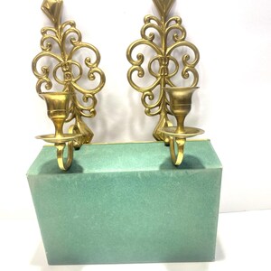Puede incluir: Dos apliques de pared dorados y ornamentados, con una parte superior en forma de diamante y detalles en espiral. Cada aplique tiene un portavelas y una peque&ntilde;a base redonda. Los apliques se muestran sobre una caja verde.
