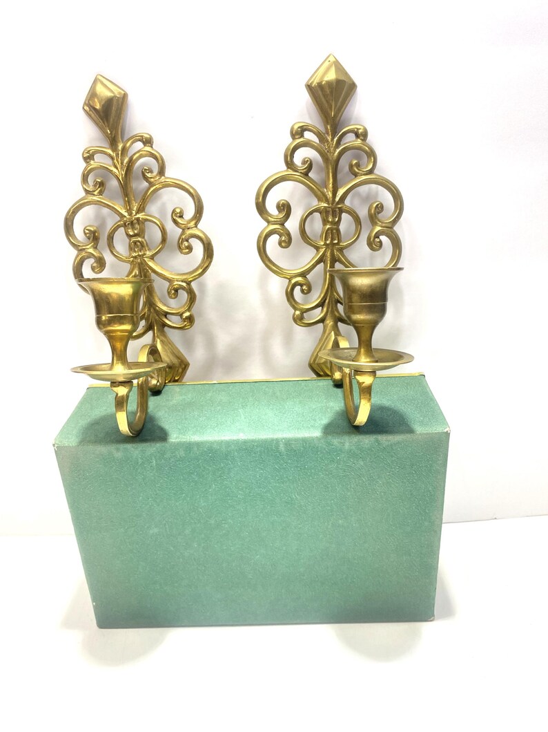 Puede incluir: Dos apliques de pared dorados y ornamentados, con una parte superior en forma de diamante y detalles en espiral. Cada aplique tiene un portavelas y una peque&ntilde;a base redonda. Los apliques se muestran sobre una caja verde.